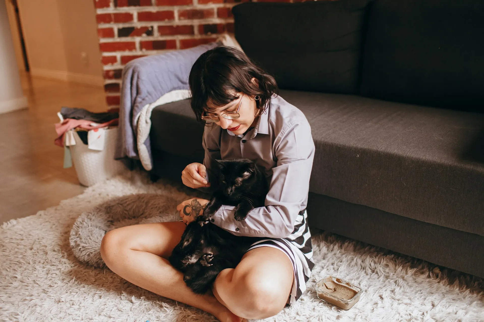 A person sits cross-legged on a fluffy white rug, gently feeding a black cat held in their arms.